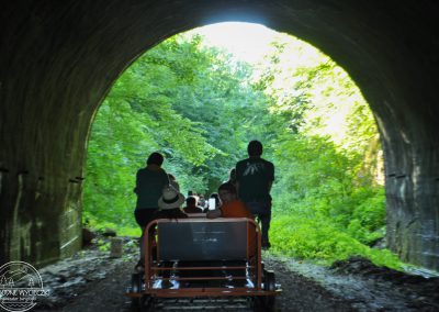Letnie zdjęcie zrobione w tunelu z widokiem na zielony las. Na torach stoi pomarańczowa drezyna a na niej siedzą cztery osoby odwrócone tyłem względem osoby oglądającej.