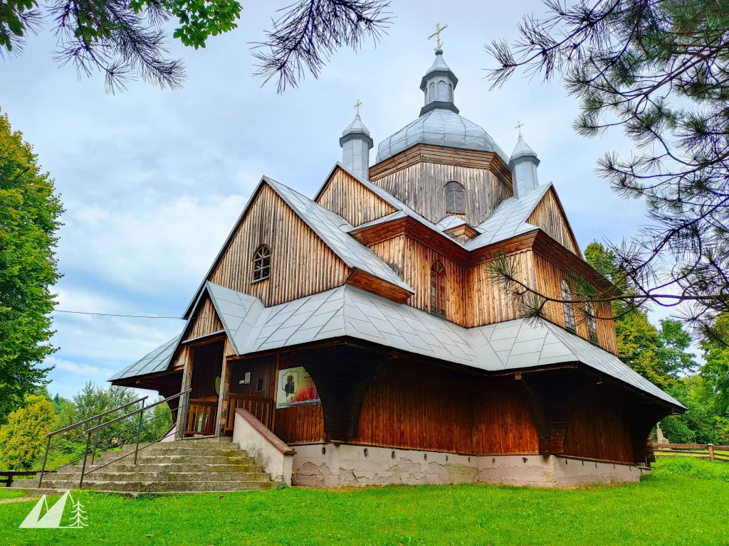 Zdjęcie przedstawiające drewnianą cerkiew, pokrytą blachą. Dach jest wielokrotnie łamany. Widać jedną główną kopułę oraz dwie znacznie mniejsze. Wszystkie są zwieńczone krzyżem.