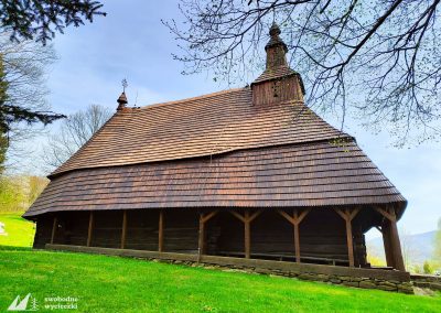 Zdjęcie przedstawia zabytkową drewnianą cerkiew. Ma prosty dach, pod którym widać coś na wzór krużganek.