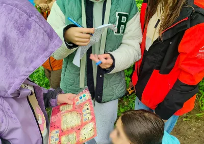 Zdjęcie przedstawia cztery dziewczyny które uczestniczą w grze terenowej w Bieszczadach. To aktywne zwiedzanie Bieszczad. z lewej strony zdjęcia stoi dziewczynka w fioletowej kurtce, z nałożonym kapturem na głowę, która w rękach trzyma kolorową kartkę z zadaniami do zrealizowania. Obok niej stoi kolejna dziewczyna w zielonej kurtce, która także ma kartkę w ręku oraz długopis. Obok niej stoi dziewczyna w czerwono - czarnej kurtce. Poniżej całej trójki kuca czwarta dziewczyna, która w ręku trzyma małą czarną skrzyneczkę. Z tyłu za nimi widać kolejnych uczestników wycieczki szkolnej w Bieszczadach.